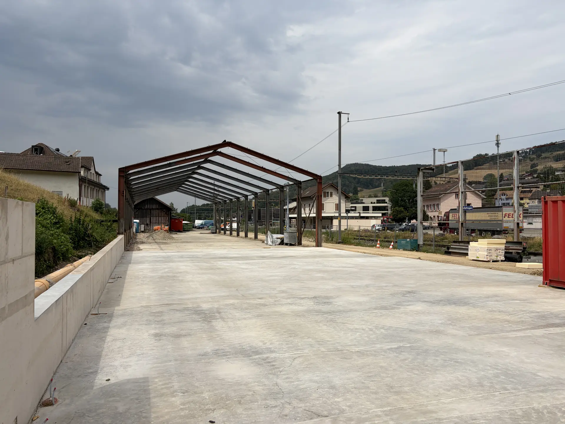 Blick der Länge nach über die neue Bodenplatte durch das Stahlskelett der Halle, im Hintergrund das Dorf