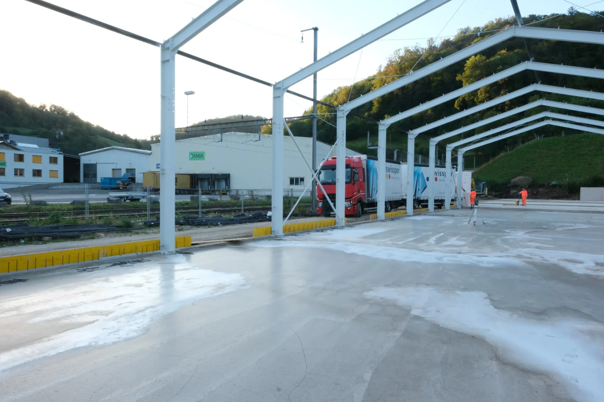 Frisch betonierte Bodenplatte der Halle unter der beschichteten Stahlkonstruktion, dahinter Bahngleise und ein Lastwagen
