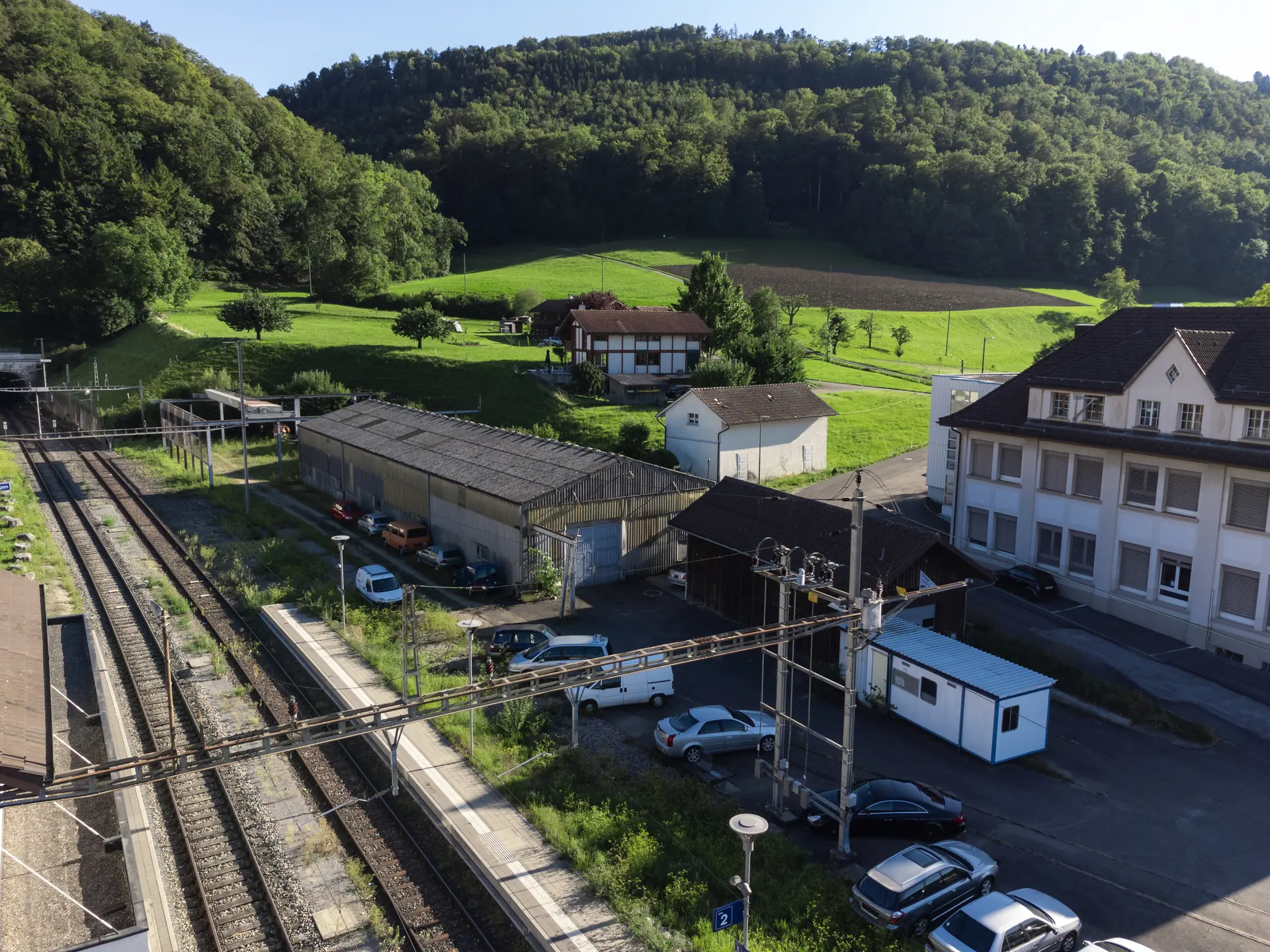 Luftbild des Areals vor dem Umbau: die Stahlbiegehalle, der hölzerne Güterschopf, dazwischen Parkplätze, daneben die Bahngleise vor dem grünen Hang