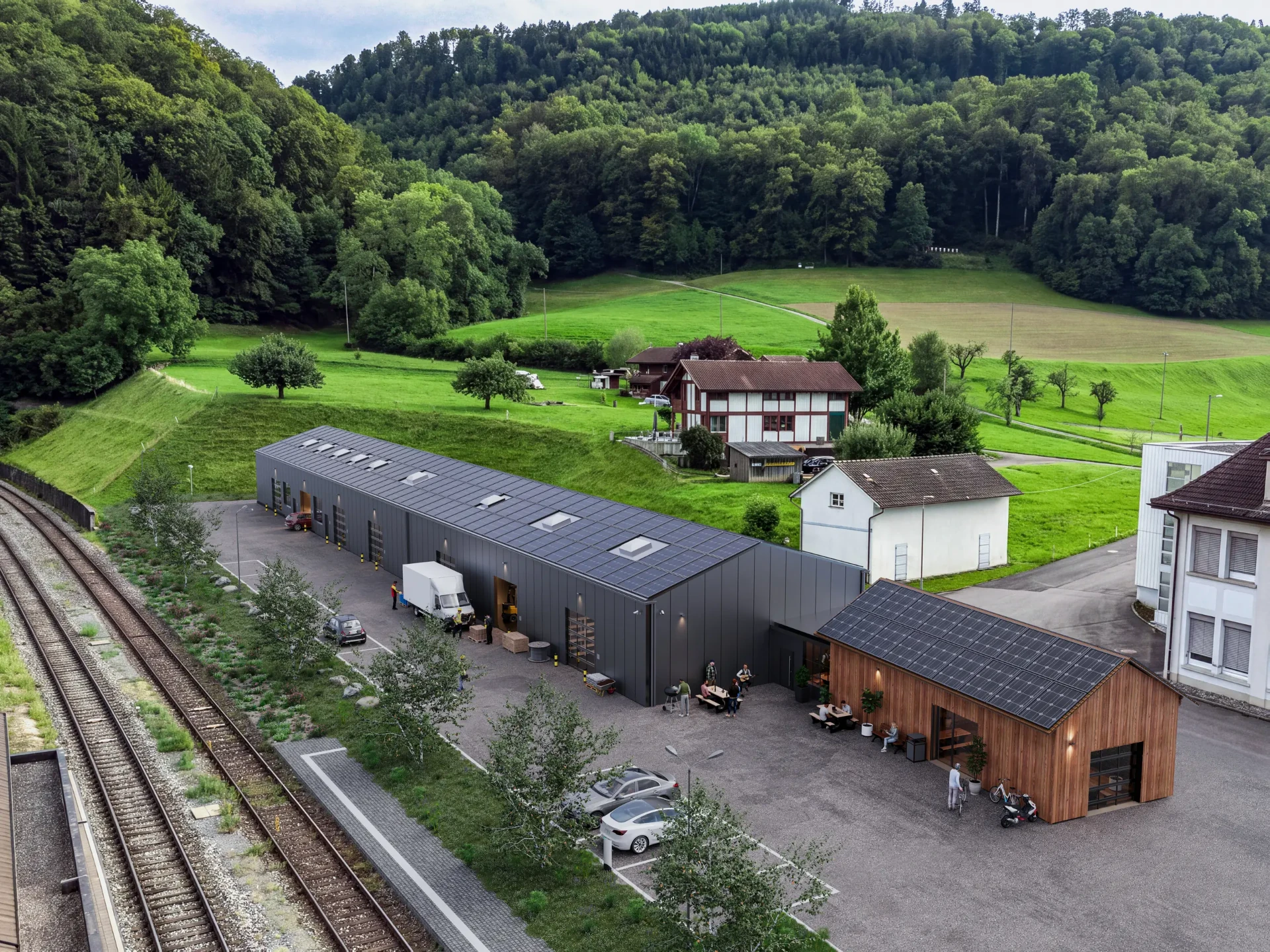 Das Areal aus der Luft: die Langhalle mit Sektionaltoren, ein Lieferwagen an einem offenen Tor, rechts der Güterschopf in Holz mit Photovoltaik, im Vordergrund die Bahngleise