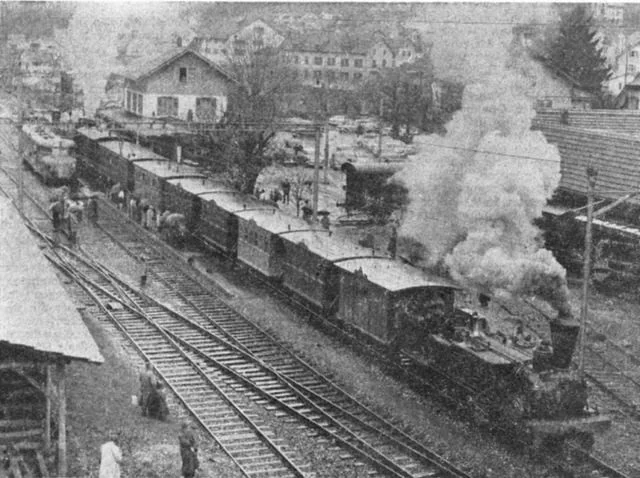 Schwarzweissaufnahme des Bahnhofs Läufelfingen von 1958: ein Dampfzug mit Güter- und Personenwagen steht an den Gleisen, dahinter das Aufnahmegebäude und das Dorf.